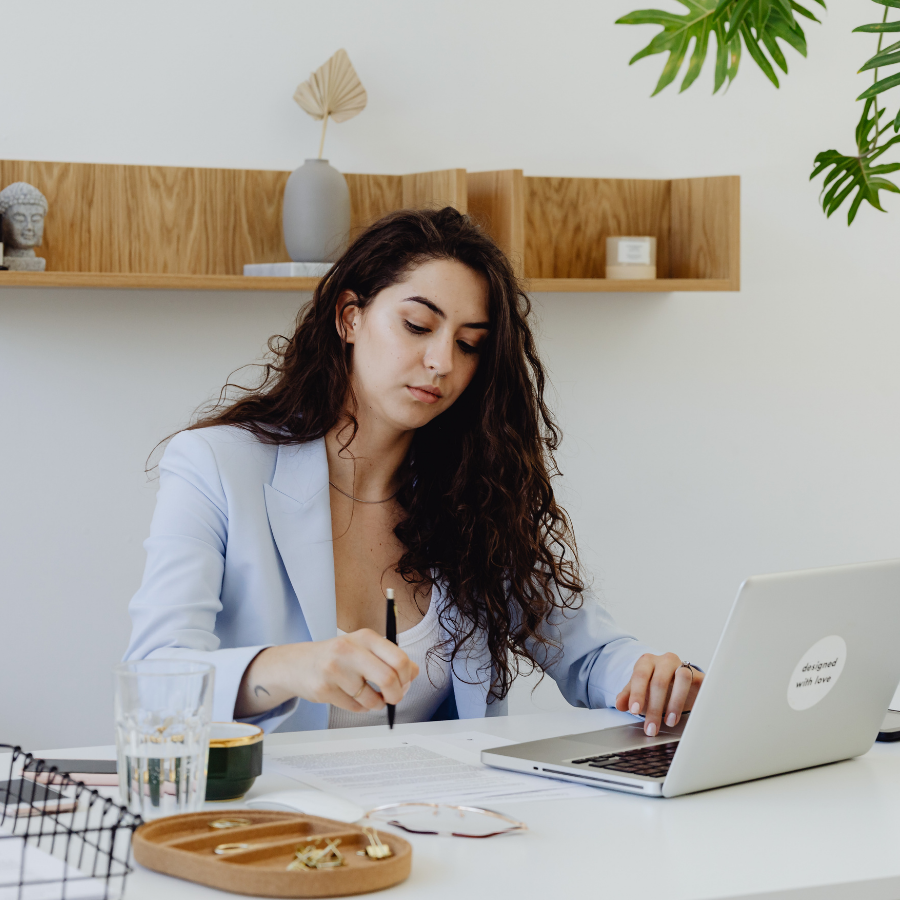 Woman with long curly hair working at a desk with a laptop, pencil, and paper in a modern, minimalistic office with plants and decorative shelves.