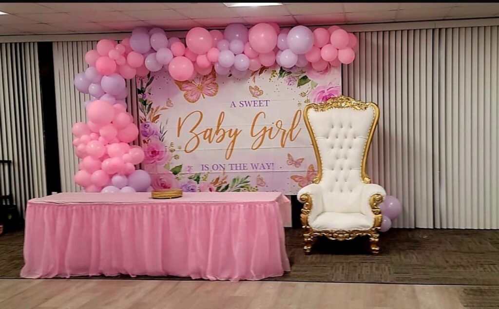 Baby shower setup with pink balloons, a decorative chair, and a "Baby Girl" banner.