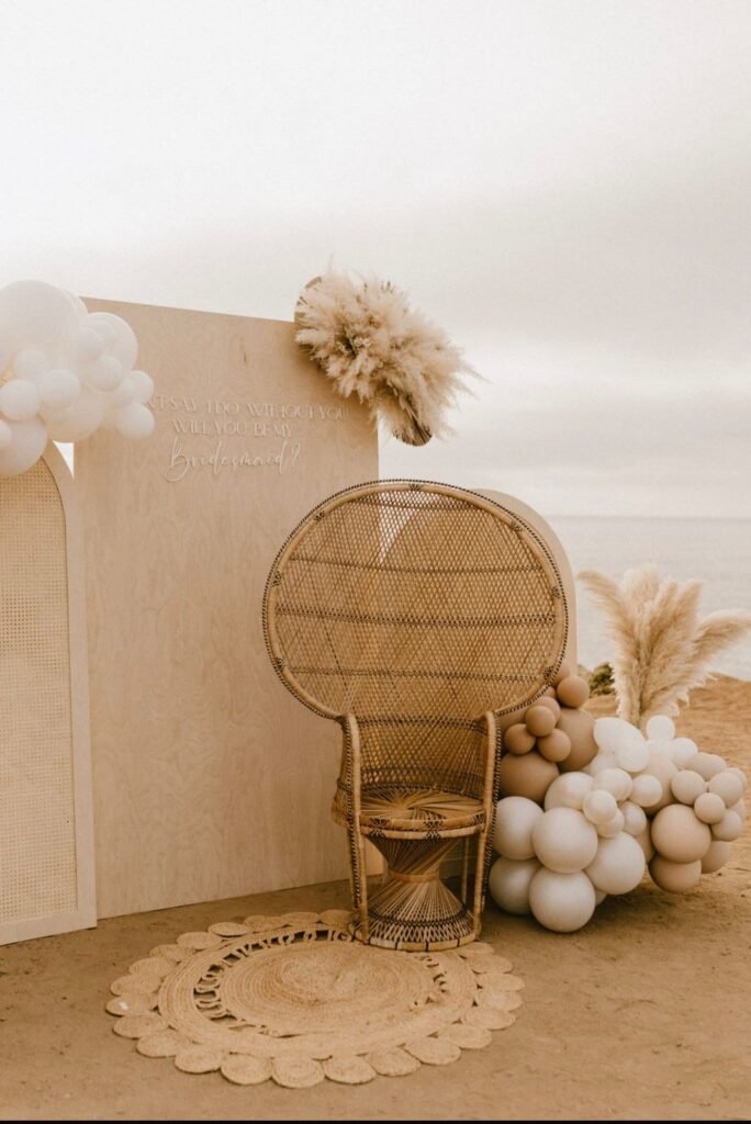 Bohemian beach setting with wicker chair, beige background, pampas grass, white and beige balloons, and decorative rug.