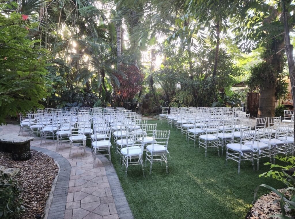 Outdoor wedding venue with arranged white chairs on grass, surrounded by lush greenery.