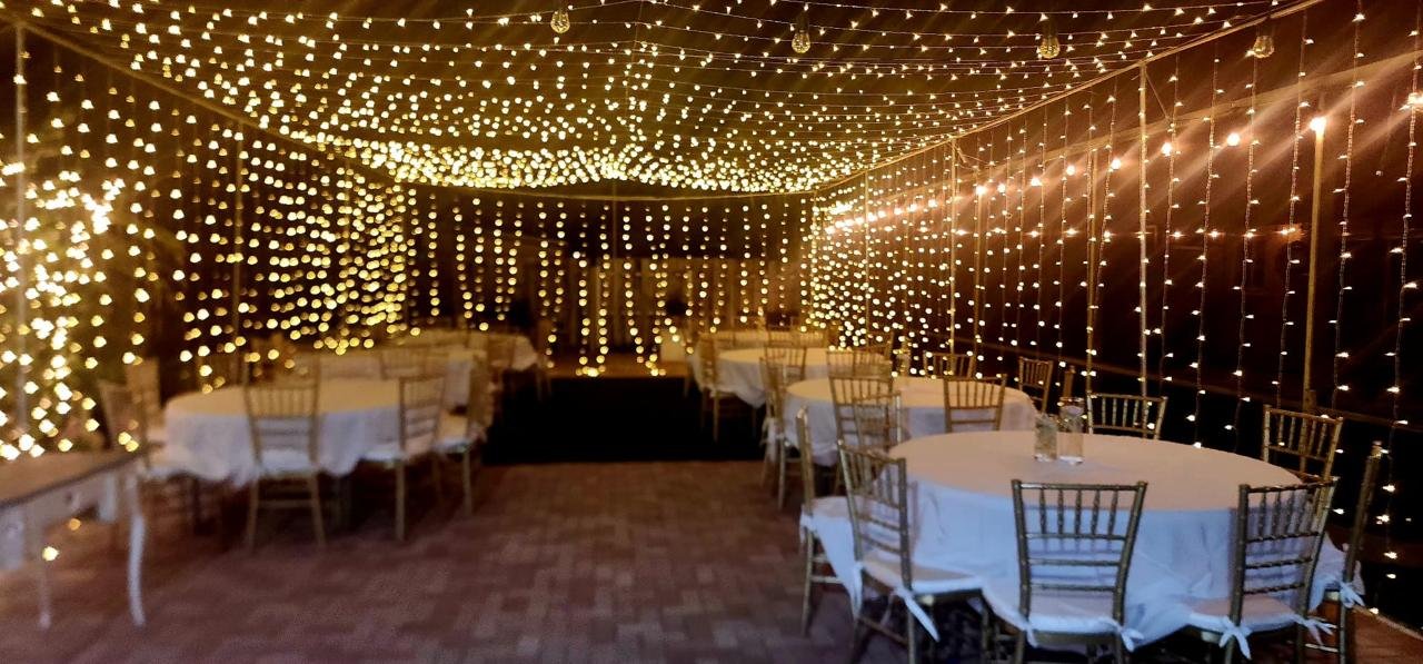 Decorative event space with hanging string lights, round tables with white cloths, and gold chairs.