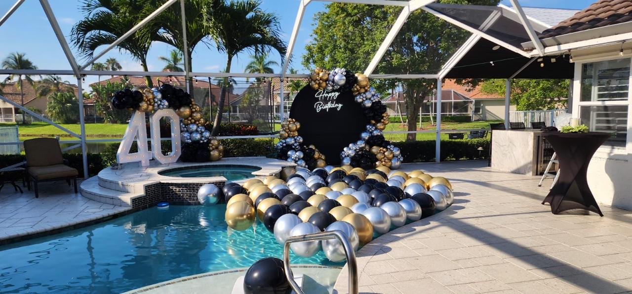 Outdoor 40th birthday party setup with a pool, black and gold balloon decorations, large "40" sign, and a "Happy Birthday" backdrop.