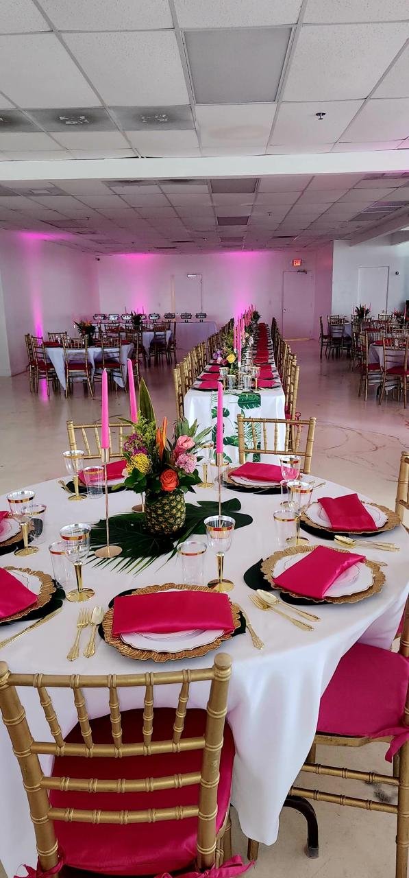 Elegant banquet hall with tables set for an event, featuring gold chairs, white tablecloths, pink napkins, and floral centerpieces with pineapples. Ambient pink lighting decorates the walls.