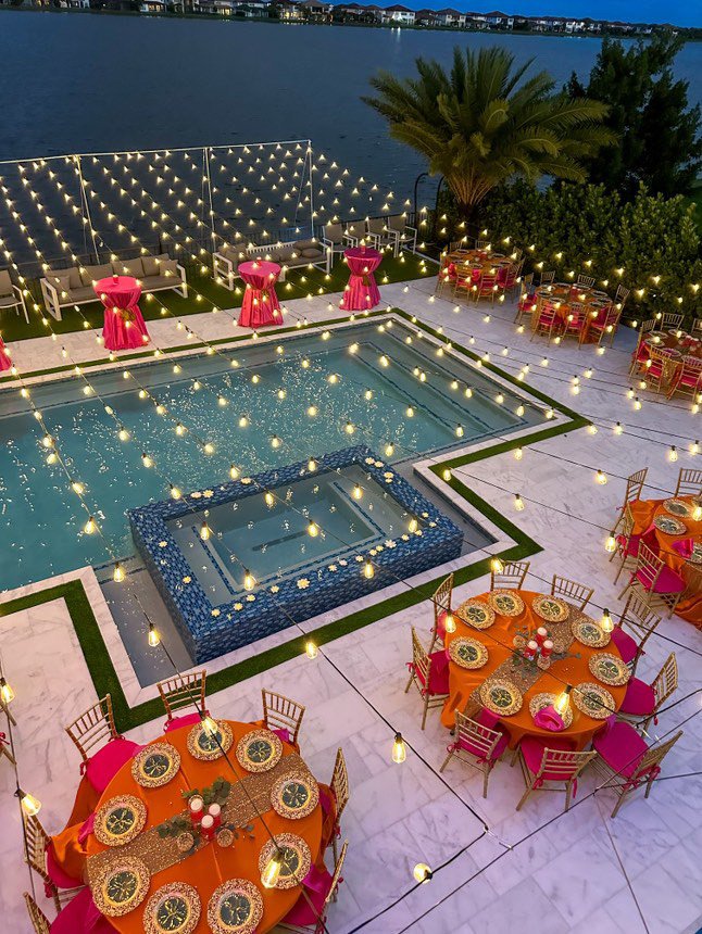 Outdoor poolside event setup with string lights. Tables with red tablecloths and gold chairs are arranged near the pool. Pink cocktail tables and outdoor seating are visible. A water view and palm trees are in the background.