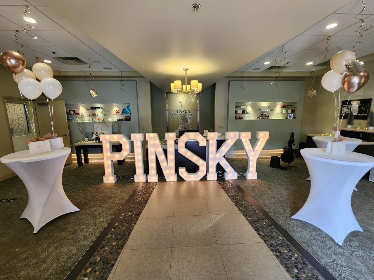 Elegant indoor setting with large illuminated letters spelling "PINSKY," flanked by white-draped tables and balloons. Festive decorations, including small gift boxes, adorn the room.