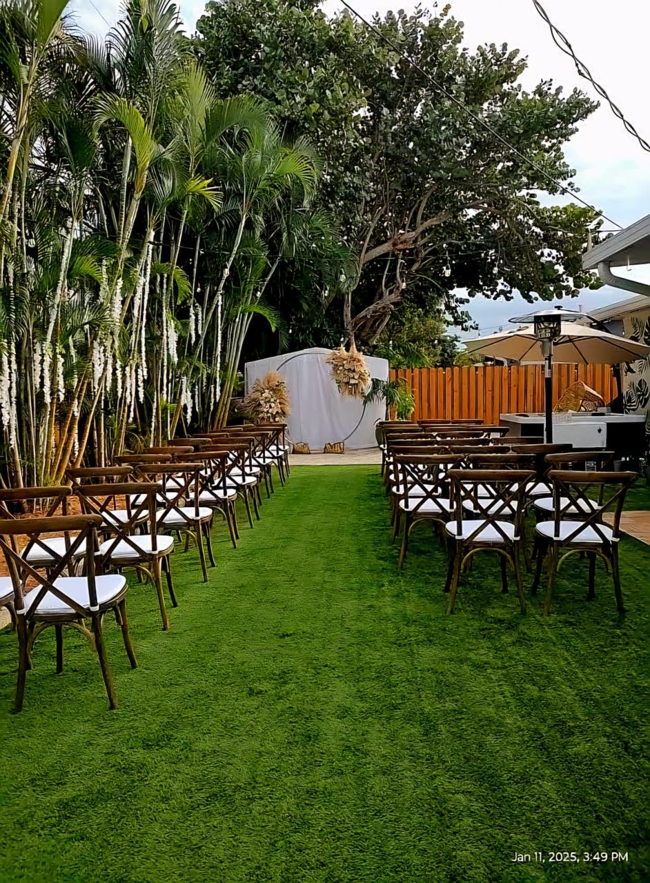 Outdoor wedding ceremony setup with wooden chairs, a grassy aisle, and a decorated arch, surrounded by palm trees and greenery.