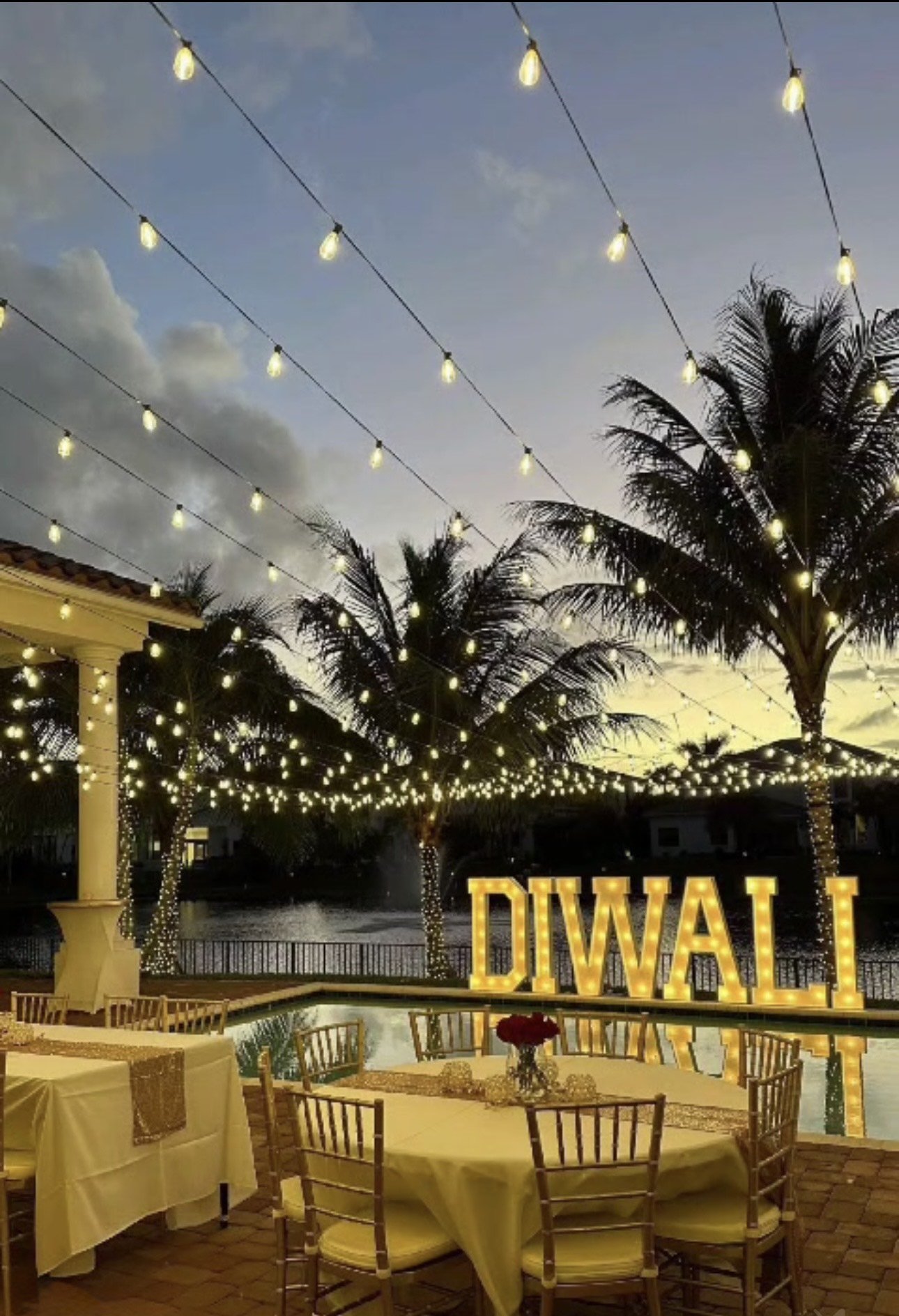 Outdoor Diwali celebration with string lights, palm trees, tables, chairs, and illuminated "DIWALI" sign by a pool.