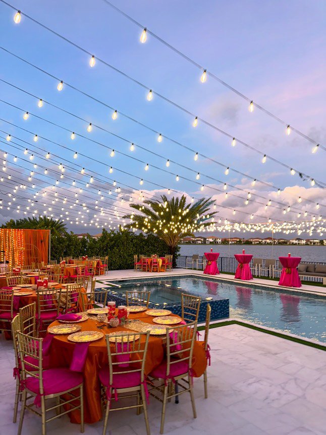Outdoor poolside event setup with string lights, pink and orange table coverings, gold chairs, and a view of a lake.