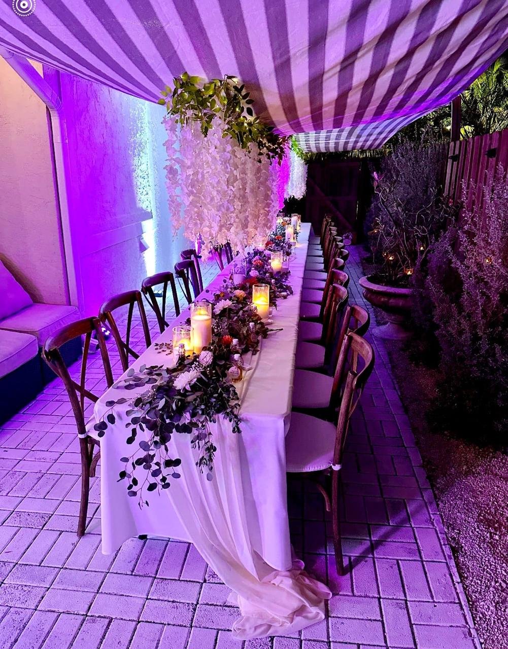 Outdoor dining table setup with candles, floral garland, and purple lighting under a striped canopy.