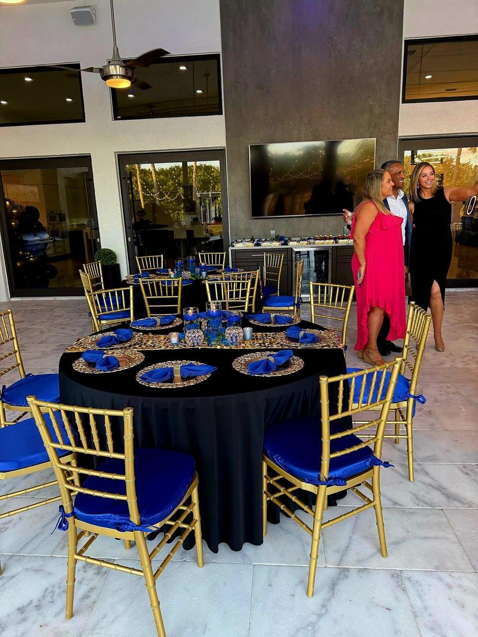 Elegant indoor event with gold chairs, a black tablecloth, blue napkins, and gold placemats on a round table. Three people are taking a selfie near a wall-mounted TV. The room has a modern design with large windows and marble flooring.