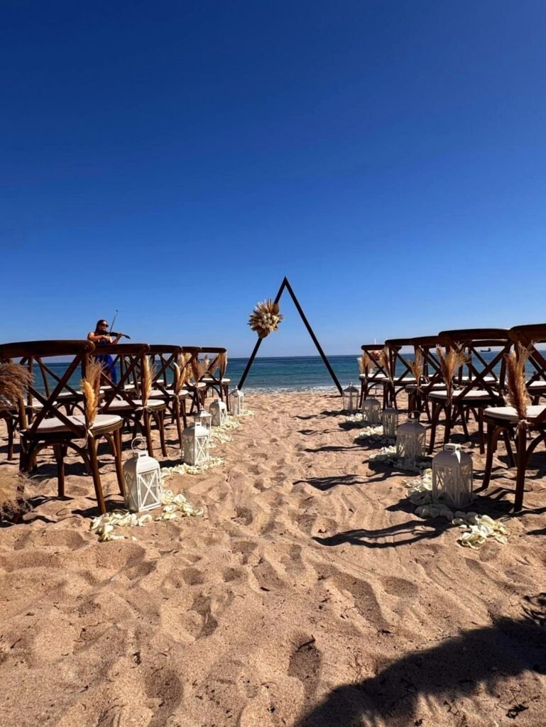 Beach wedding ceremony setup with chairs, lanterns, and a triangular arch facing the ocean. Blue sky and sand visible.