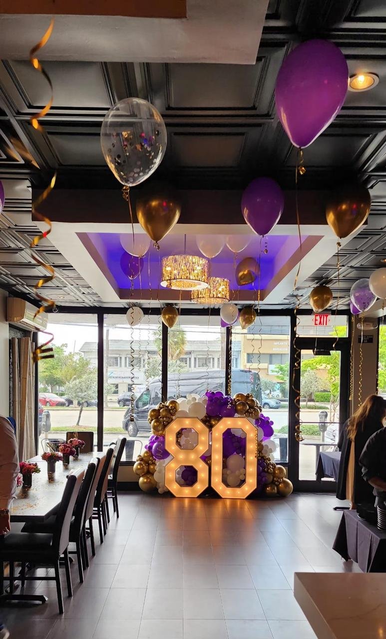 Indoor party setup with purple, white, and gold balloons and illuminated '80' decoration.