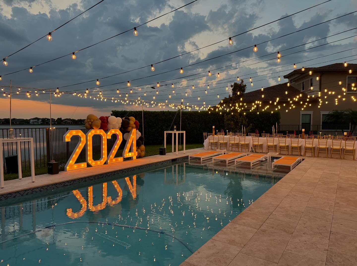A poolside event setup with illuminated "2024" sign, string lights overhead, a decorated table with chairs, and a backdrop of a lake and residential buildings at sunset.