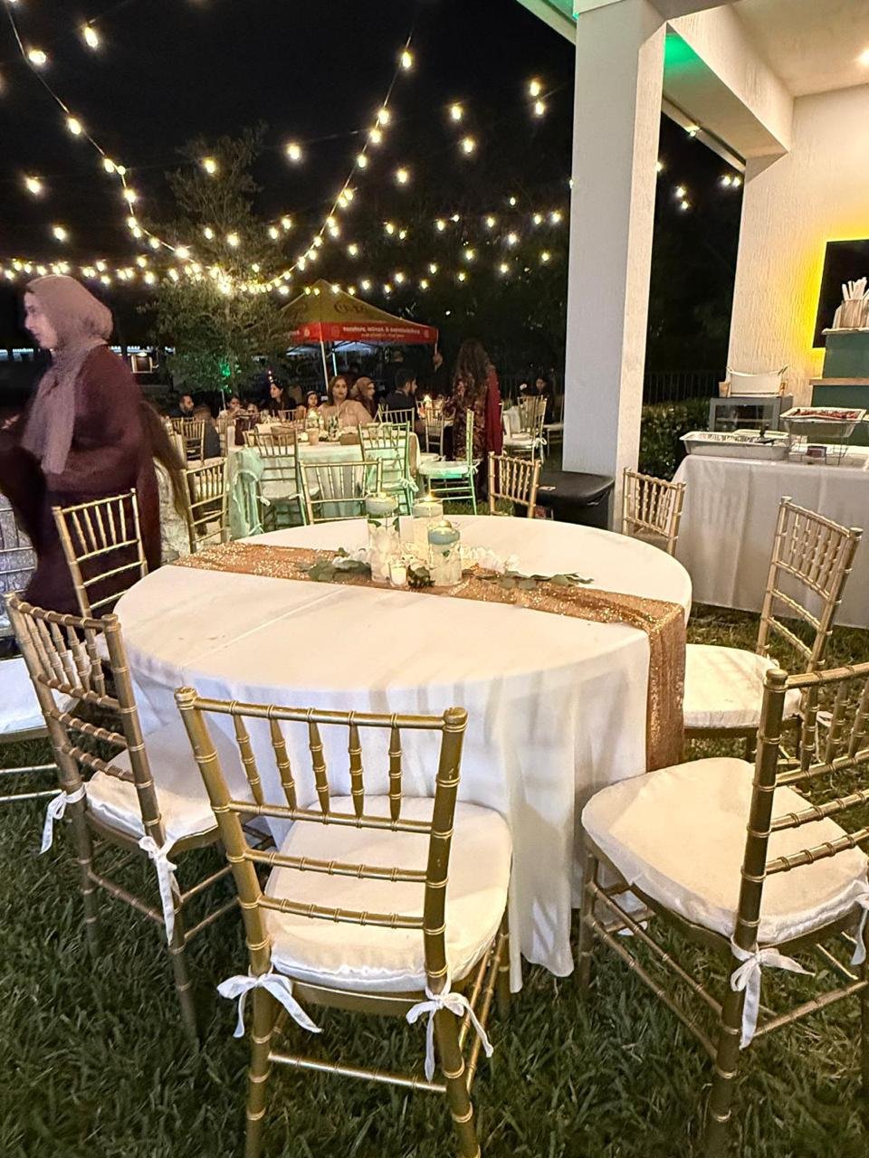 Outdoor wedding reception setup at night with string lights, decorated tables, gold chairs, and attendees. The scene is festive and elegant.