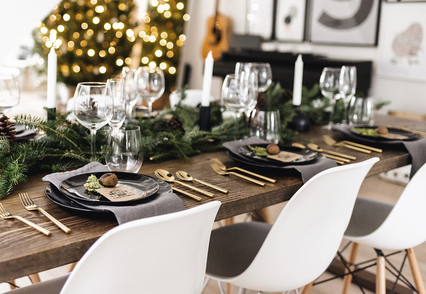 Festive dining table setting with greenery, candles, and gold utensils, against a backdrop of a decorated Christmas tree.
