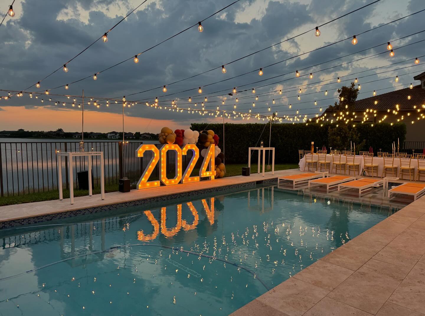 Poolside party setup with large lit "2024" sign, string lights overhead, decorated tables, and colorful balloons at sunset.
