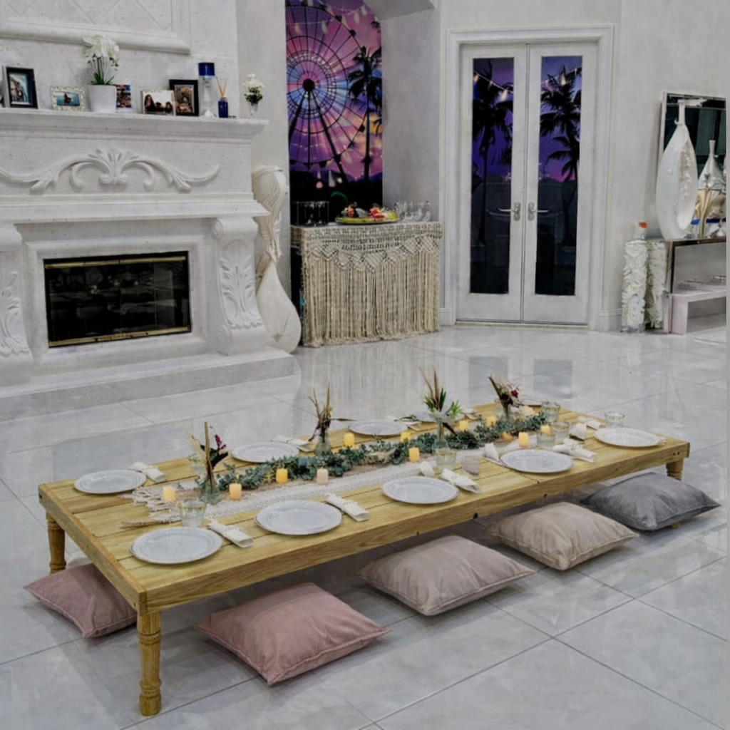 A low dining table set for a meal with light pink velvet cushions around it. The table is covered with a beige tablecloth, decorated with pampas grass centerpieces, pink napkins, and woven placemats. The room features wood cabinets, a large wall clock, and a fireplace, creating a cozy dining atmosphere.