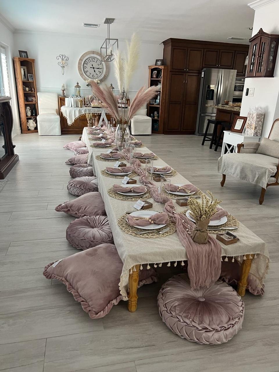 A stylish dining area featuring a low wooden table set with plates, glasses, and pink napkins. The table is adorned with pampas grass centerpieces and draped with a light beige tablecloth. Dusty pink floor cushions are arranged around the table for s