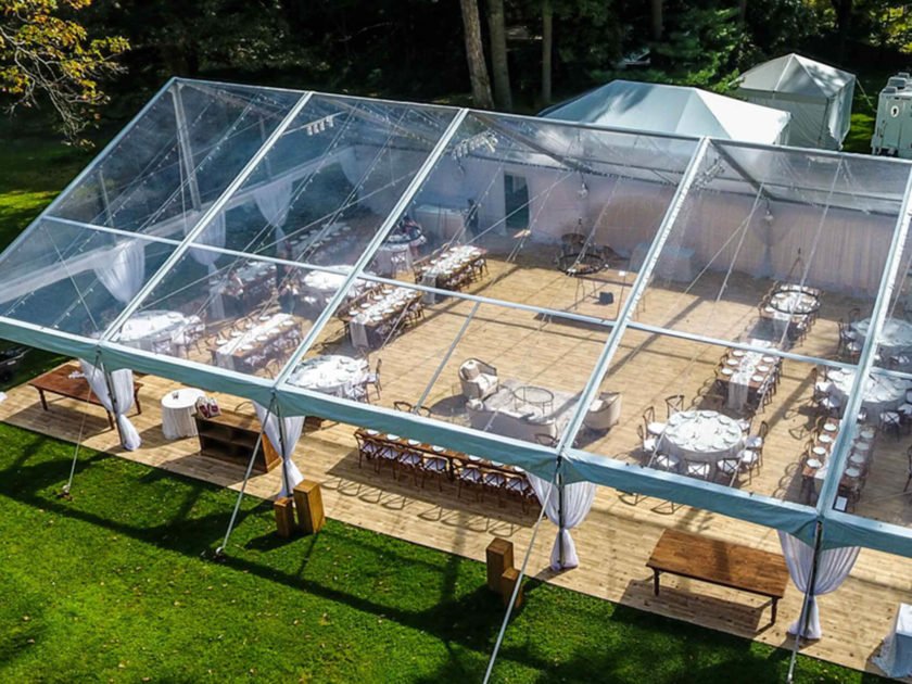 Outdoor wedding reception setup with transparent tent, tables, and chairs on a landscaped lawn.