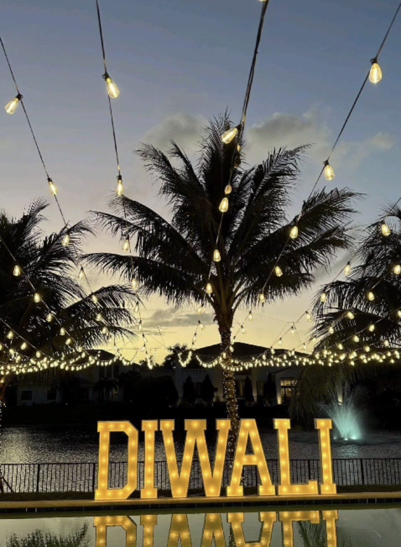 Outdoor Diwali decoration with illuminated string lights, palm trees, and large lit Diwali sign by a pool at twilight.