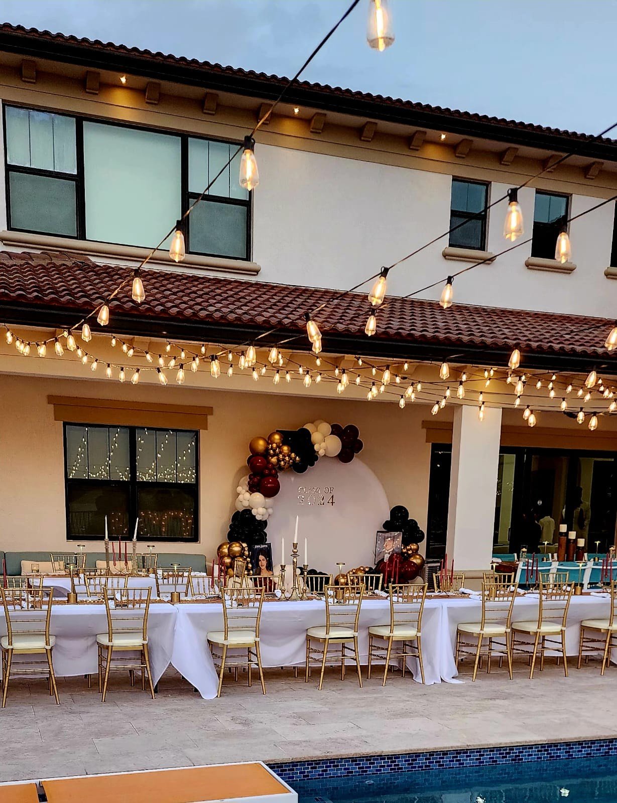 Backyard setup with a long dining table covered in white cloth and surrounded by gold chairs, set up for an outdoor event. Overhead string lights provide illumination, and a decorative arch with balloons in gold, black, and white colors is in the bac