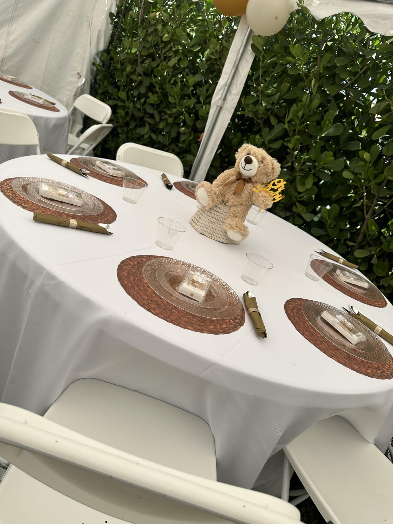 Outdoor event table setup with white tablecloth, wicker placemats, and folded napkins. A teddy bear centerpiece is on the table, surrounded by brown and white balloons. White folding chairs are arranged around the table.