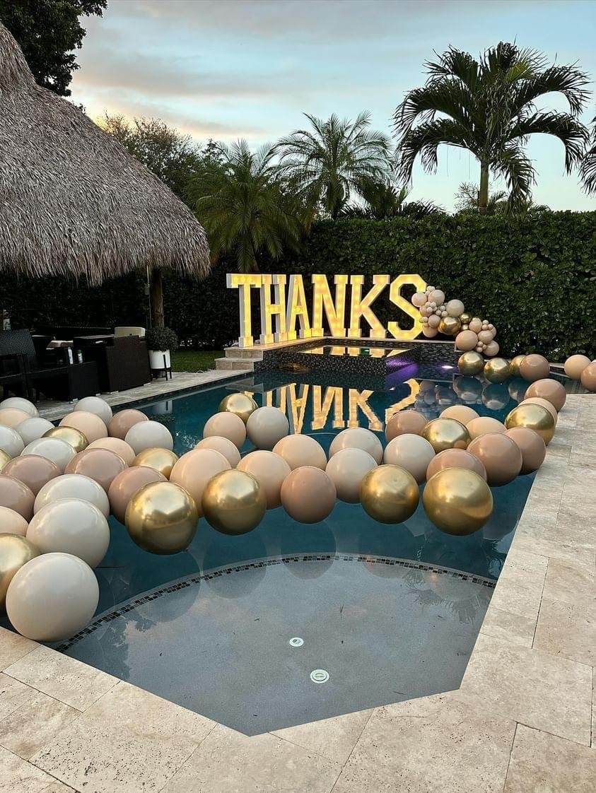 Poolside decoration for a celebratory event featuring large "THANKS" sign and clusters of gold and beige balloons, surrounded by tropical foliage and straw-roofed structure.