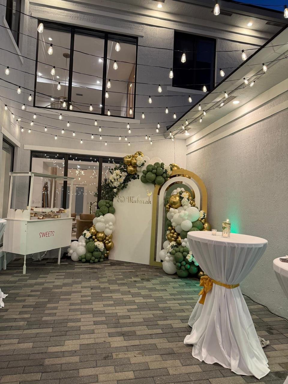 Outdoor celebration setup with string lights, decorated with green and gold balloons and flowers. A round table draped in white cloth with a gold ribbon, and a cart labeled "sweets" are visible. Backdrop says "Eid Mubarak."