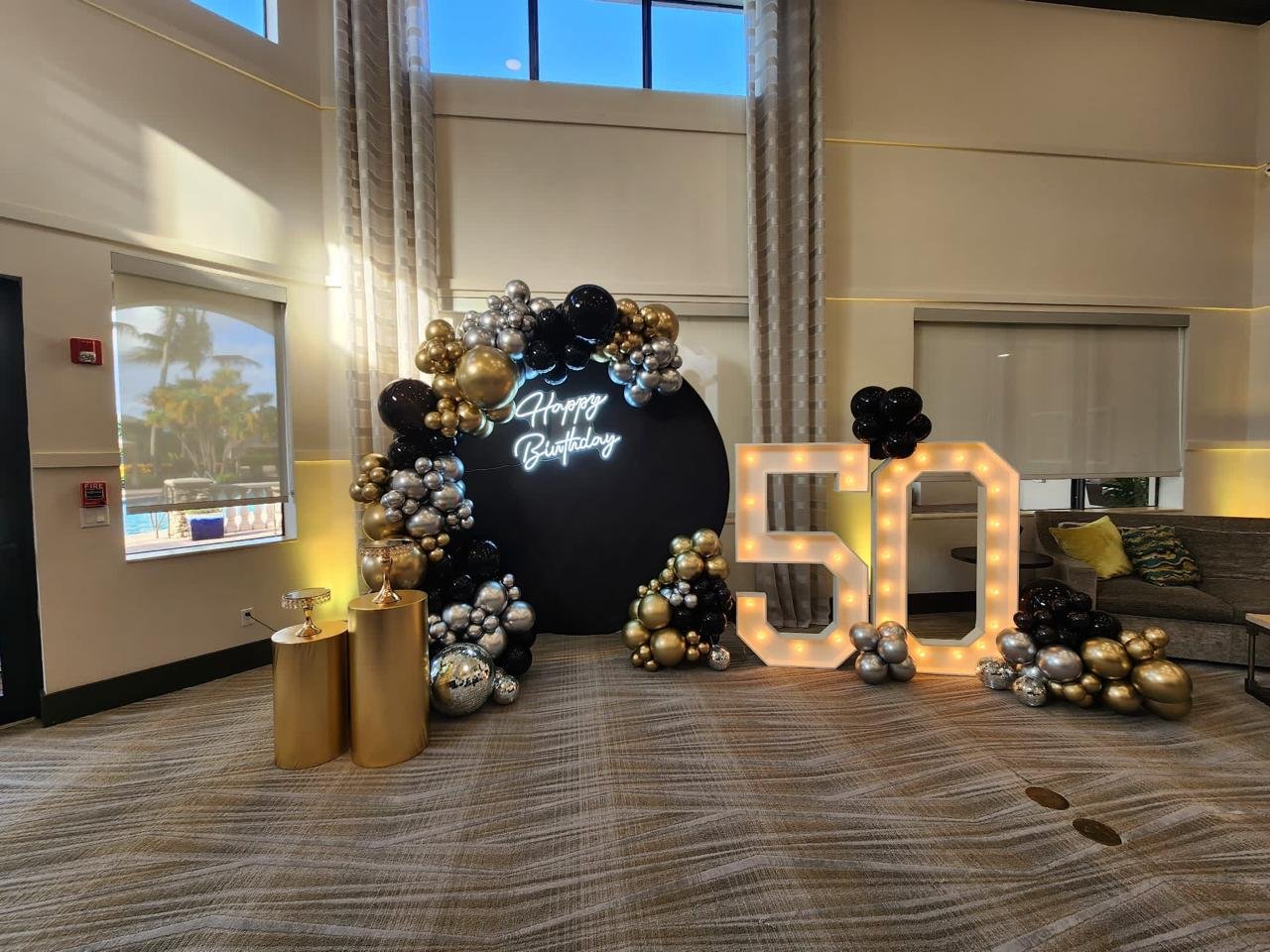 Decorative setup for a 50th birthday celebration with black, gold, and silver balloons, a "Happy Birthday" neon sign, and illuminated "50" numbers.
