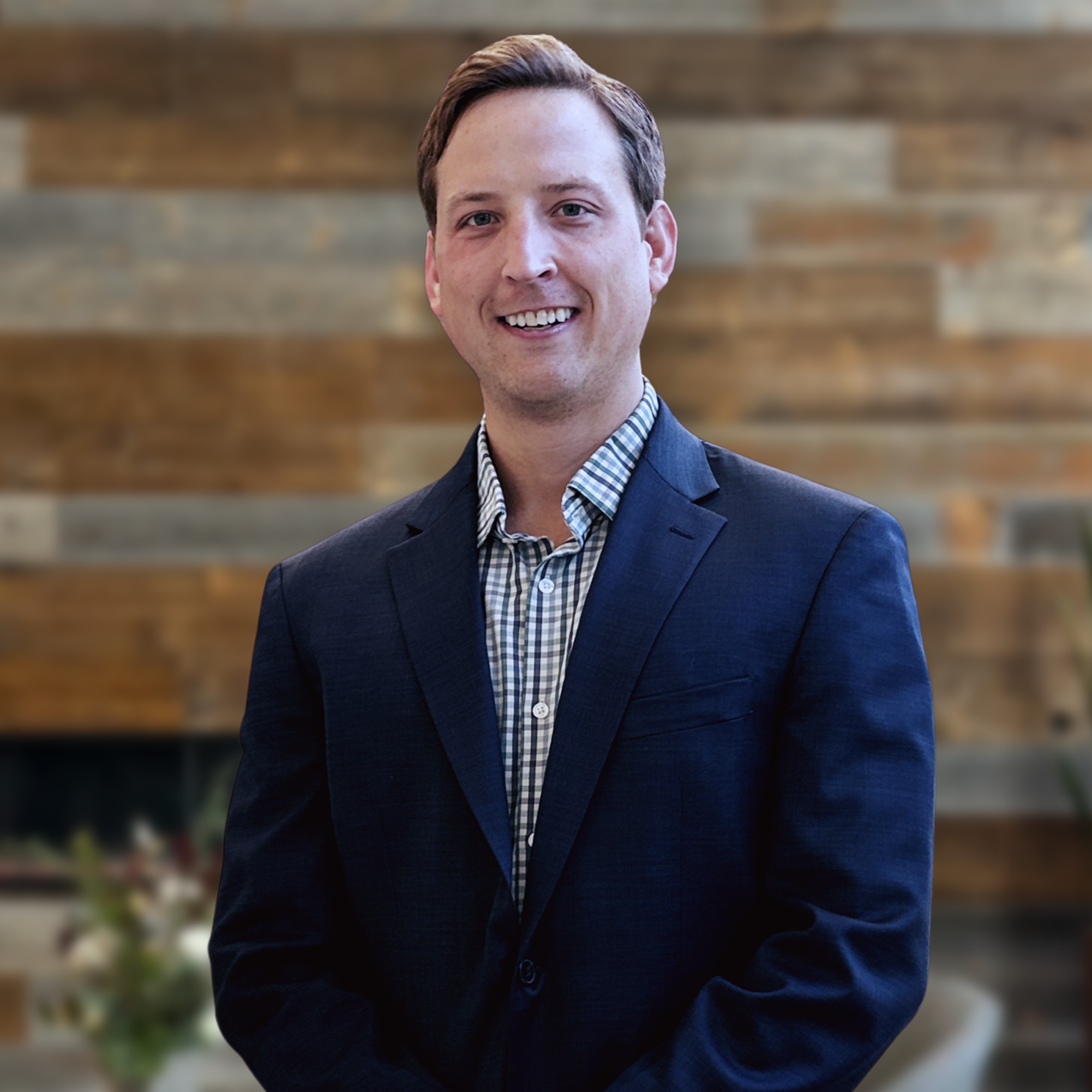 A young man with light brown hair and light skin, smiling, dressed in a dark blazer and checkered shirt, standing in front of a wooden wall background.