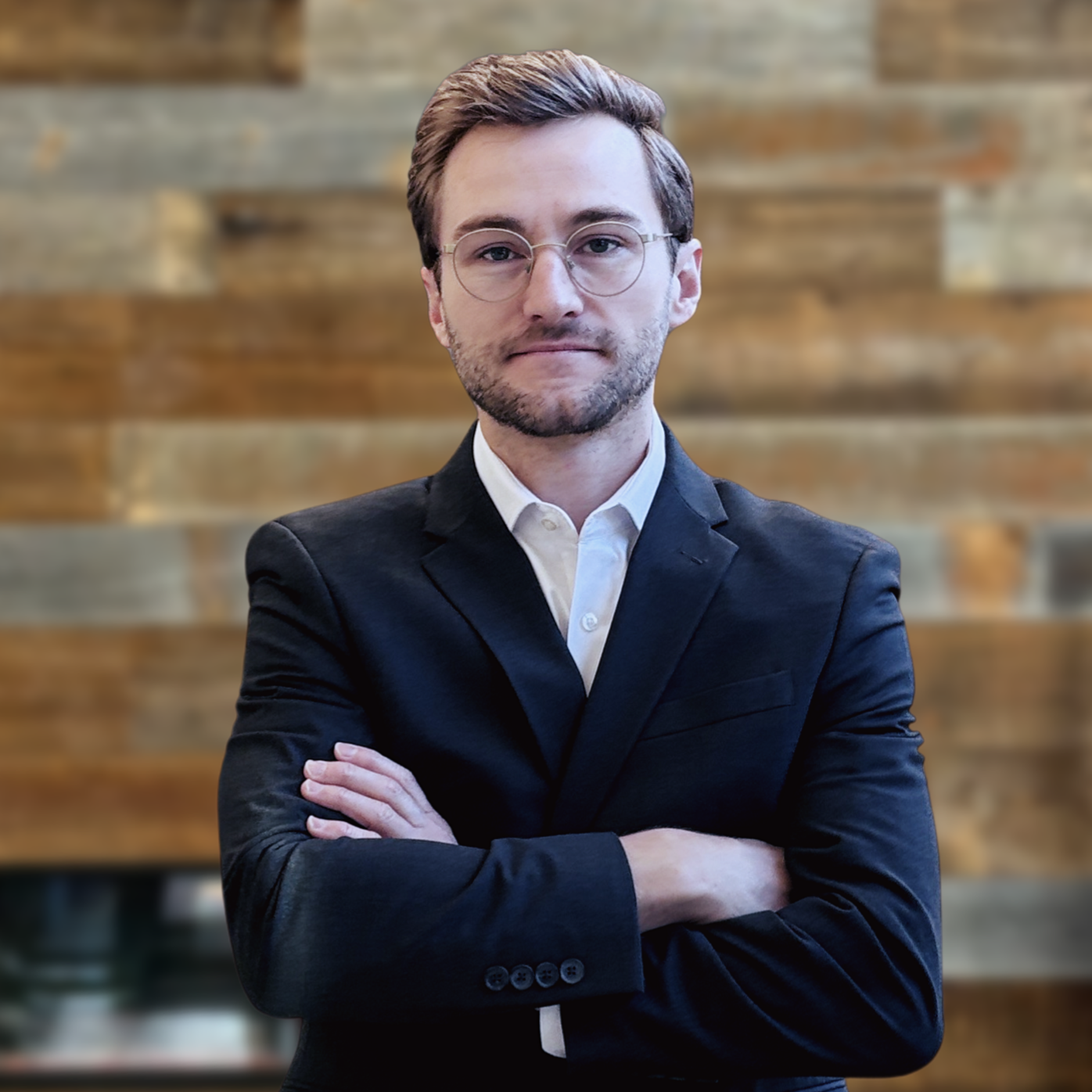 A man with brown hair, glasses, and a beard, wearing a black suit and white shirt, standing with arms crossed in front of a wooden wall.