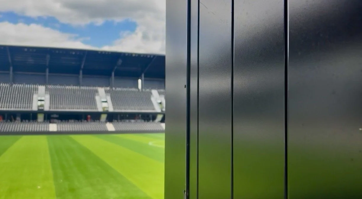 View of Ruoff Mortgage Stadium with a green field, seating area, and blue sky, seen through a side opening with a metallic wall.