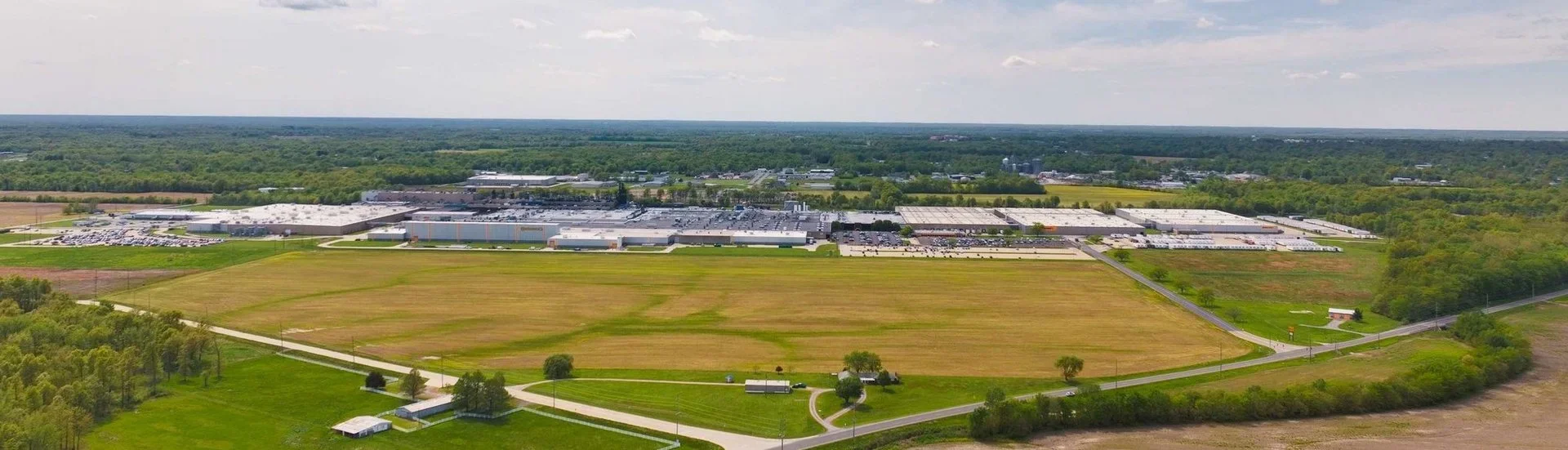Aerial view of a large industrial complex or warehouse surrounded by farmland and greenery, with roads connecting the buildings.
