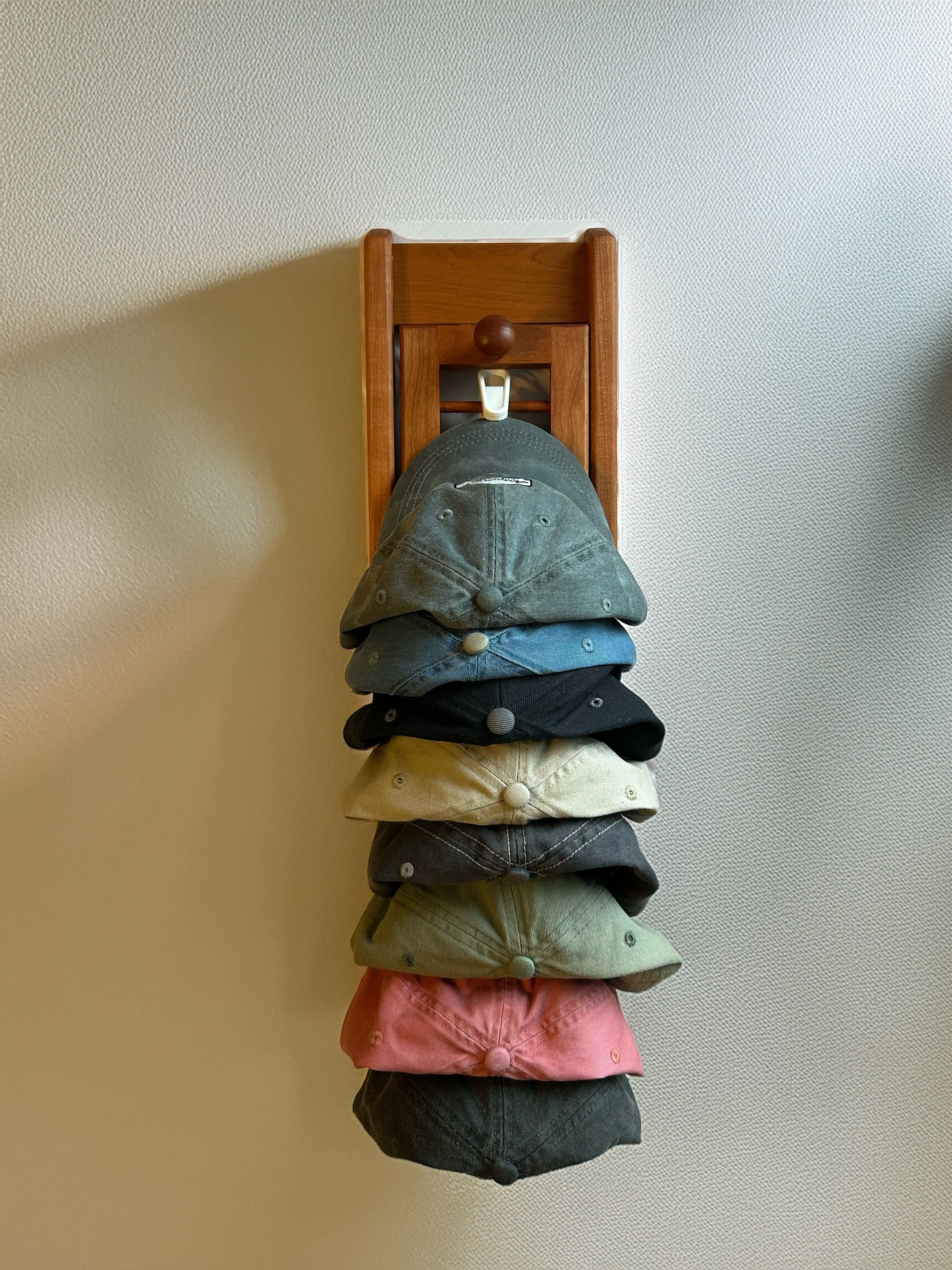 A rack of eight baseball caps hanging vertically on a wall with a wooden hook.