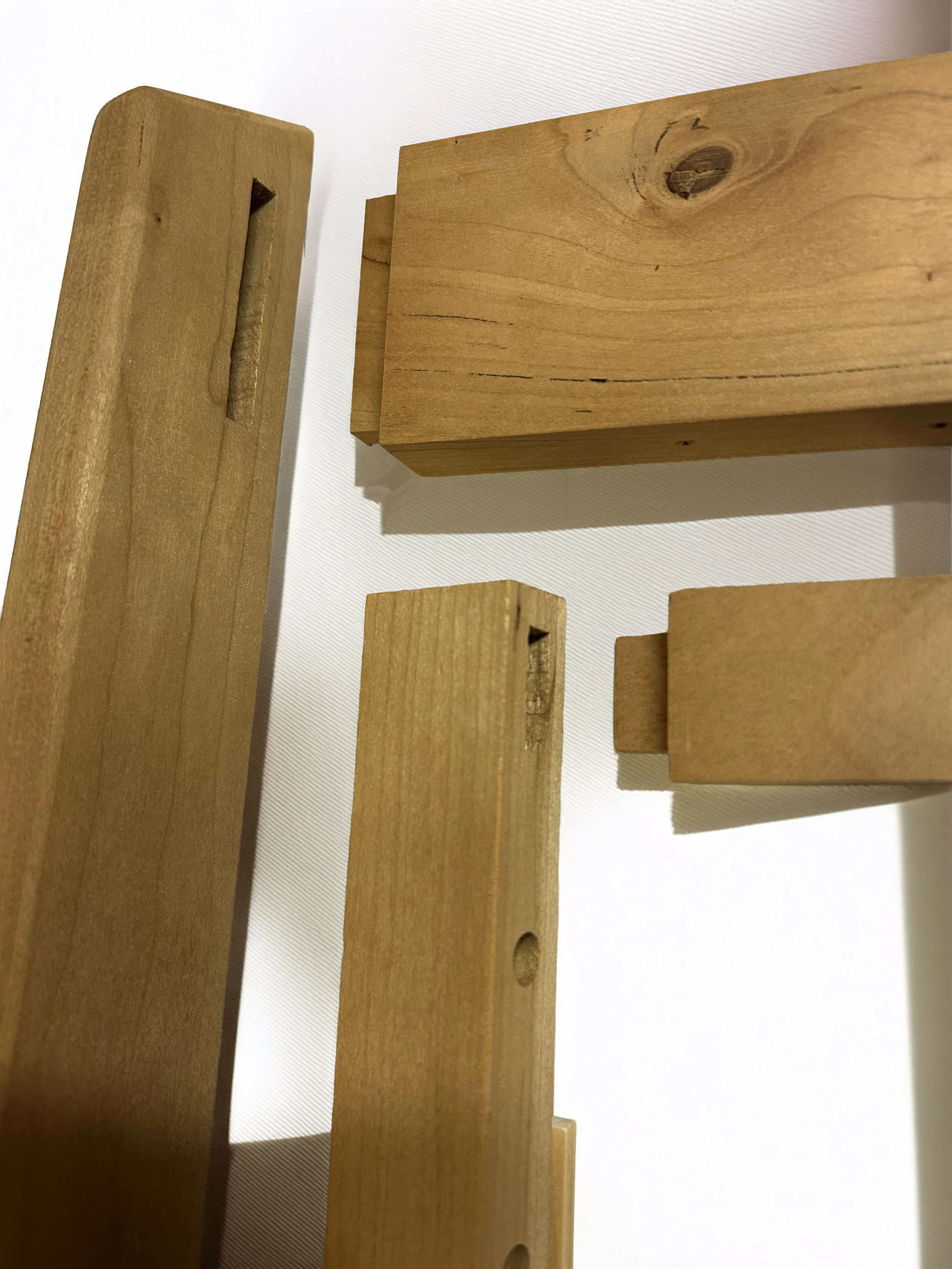 Close-up of four unfinished wooden pieces, including a rectangular beam with a knot, a flat rectangular piece, and two narrower rectangular pieces, laid on a white surface.