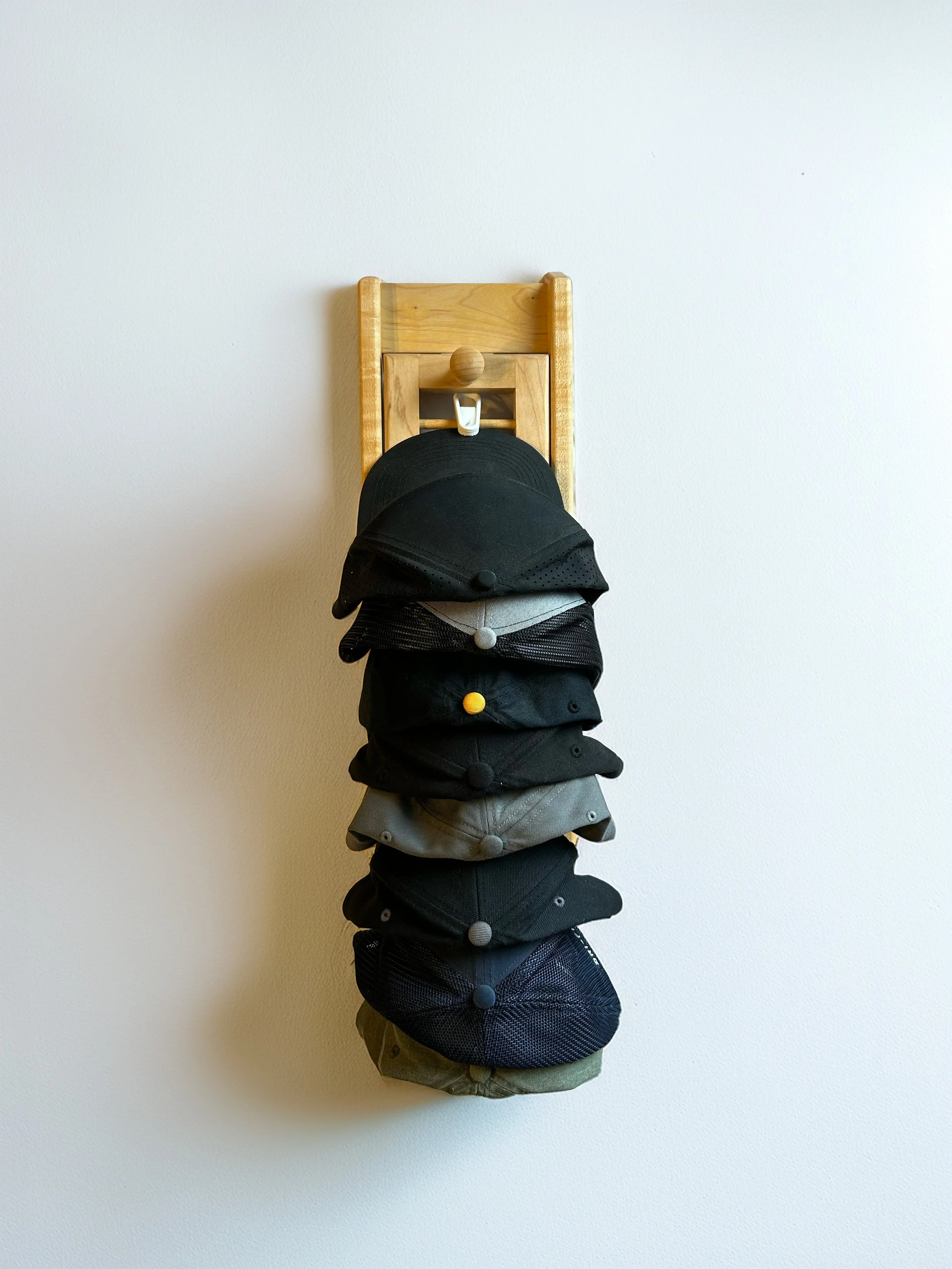 Multiple black, grey, and navy baseball caps hanging on a wooden rack mounted on a plain white wall.