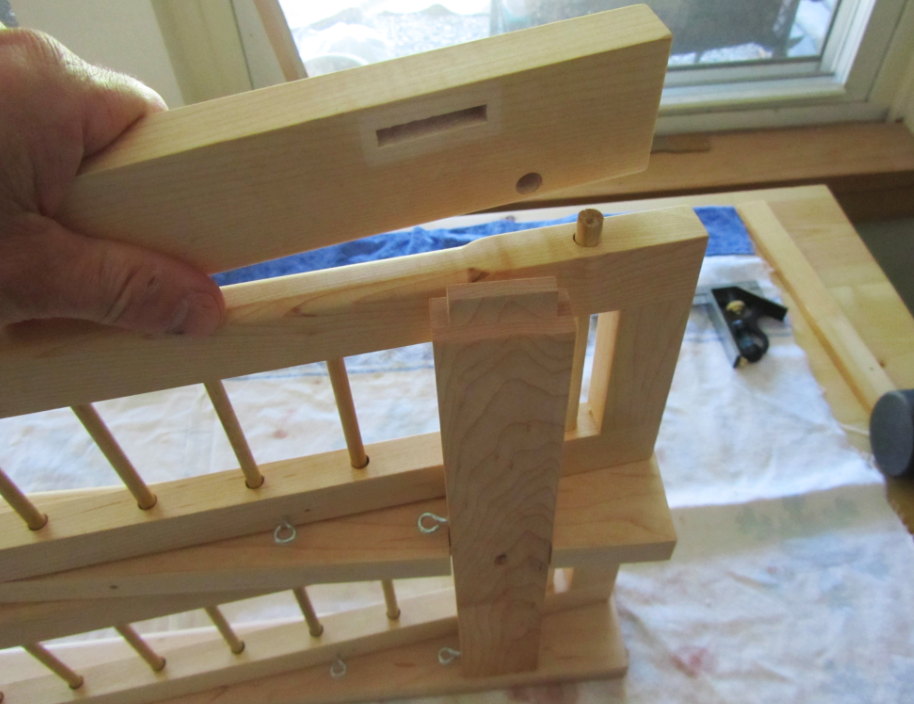 Close-up of a person's hand holding a wooden piece in a woodworking project. The project includes wooden spindles, a square cutout, and various wooden components assembled on a workbench near a window.