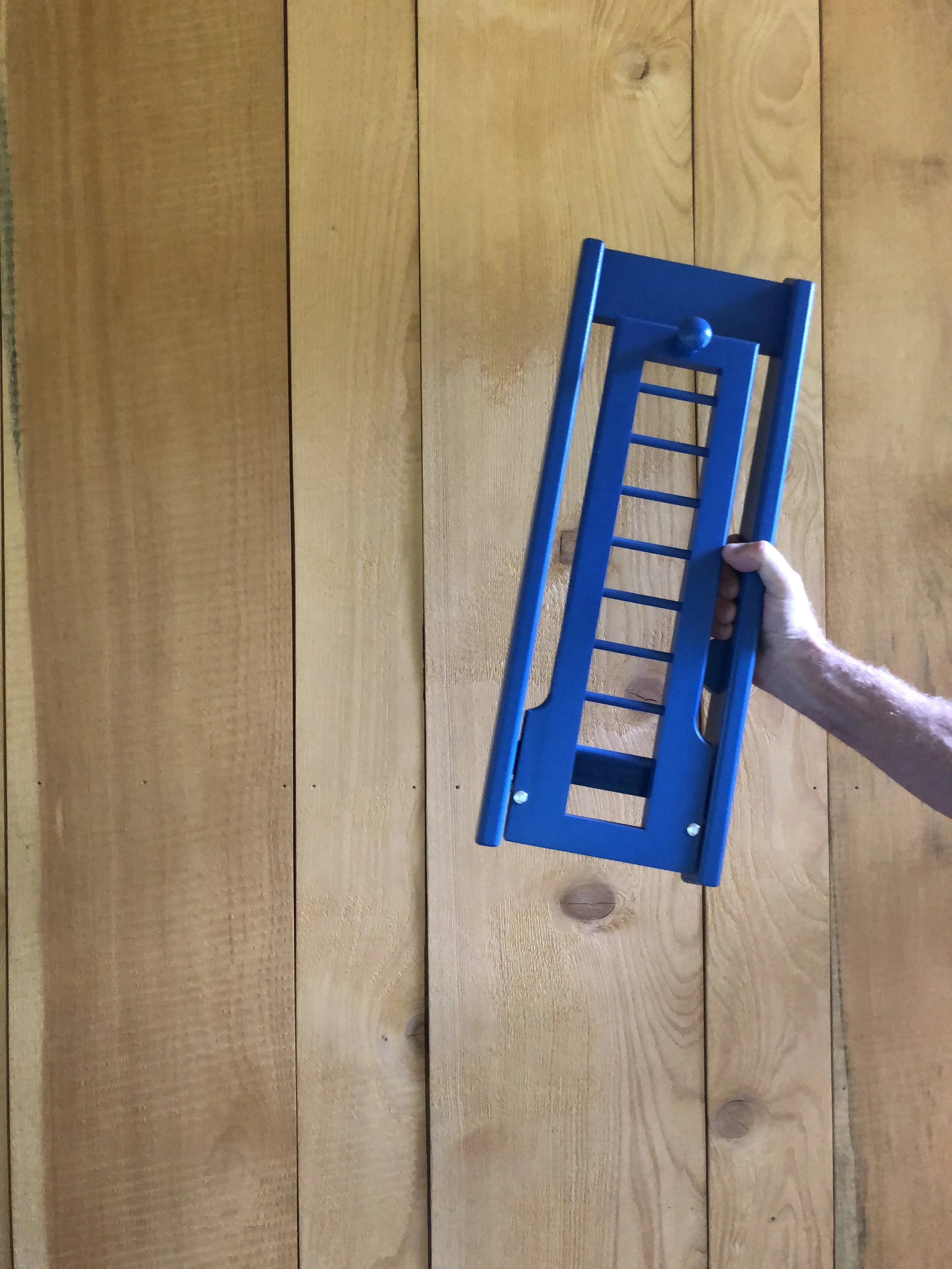 A hand holding a blue wooden hat rack against a wooden wall.