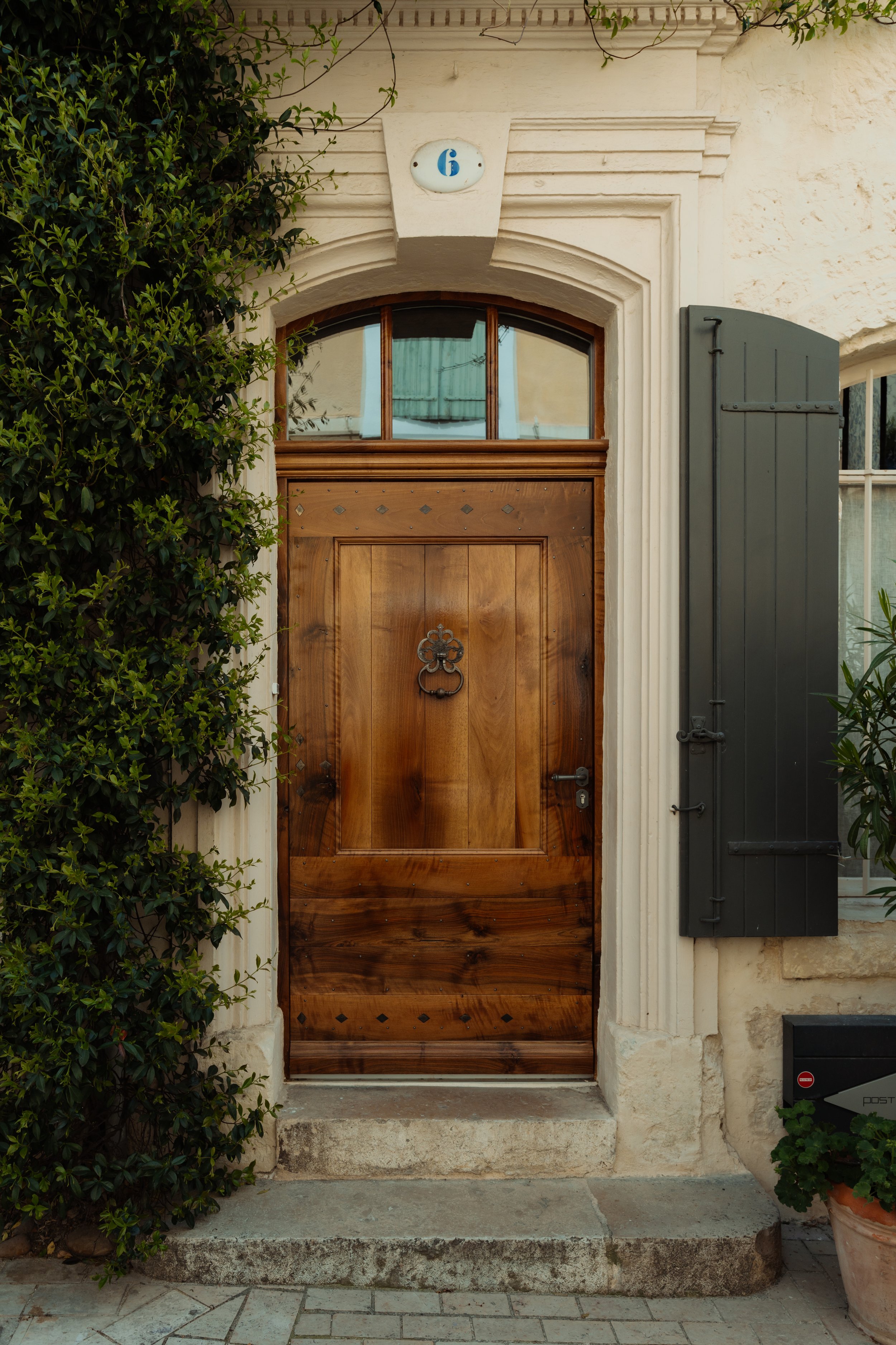 Porte en bois avec heurtoir en métal, entourée de murs en pierre et vigne grimpante, volet gris et boîte aux lettres à droite.