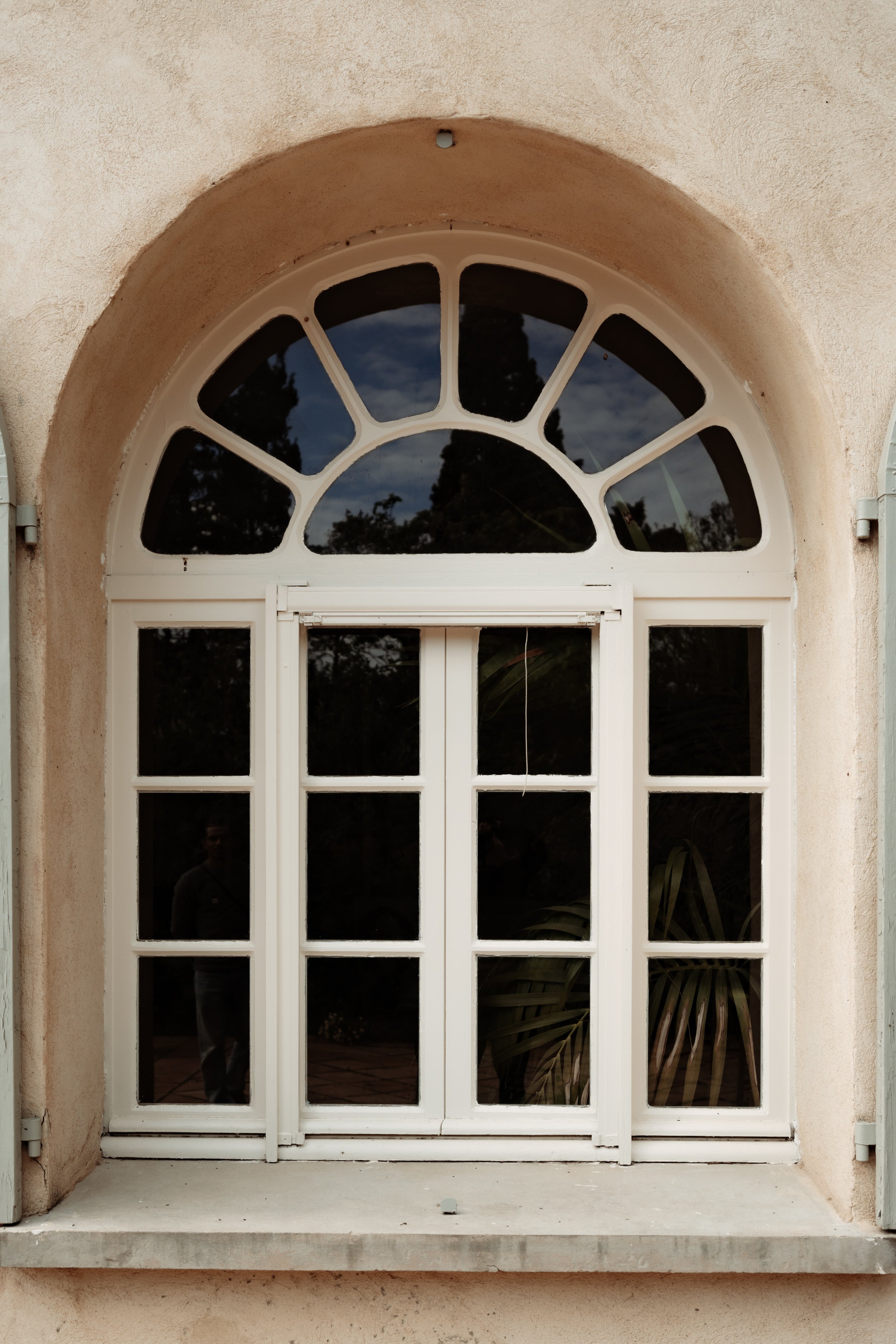 Fenêtre arquée avec cadre blanc sur un mur beige, reflet d'arbres et de feuillage visible dans le verre sombre.