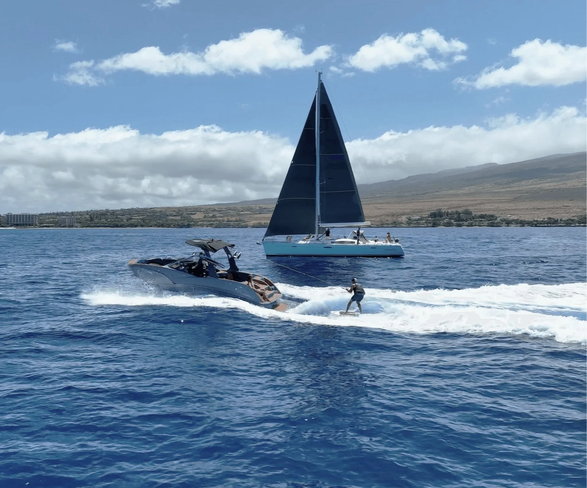 A person wakeboarding behind a boat on the ocean, with a sailboat in the background and a distant landmass with some buildings under a partly cloudy sky.