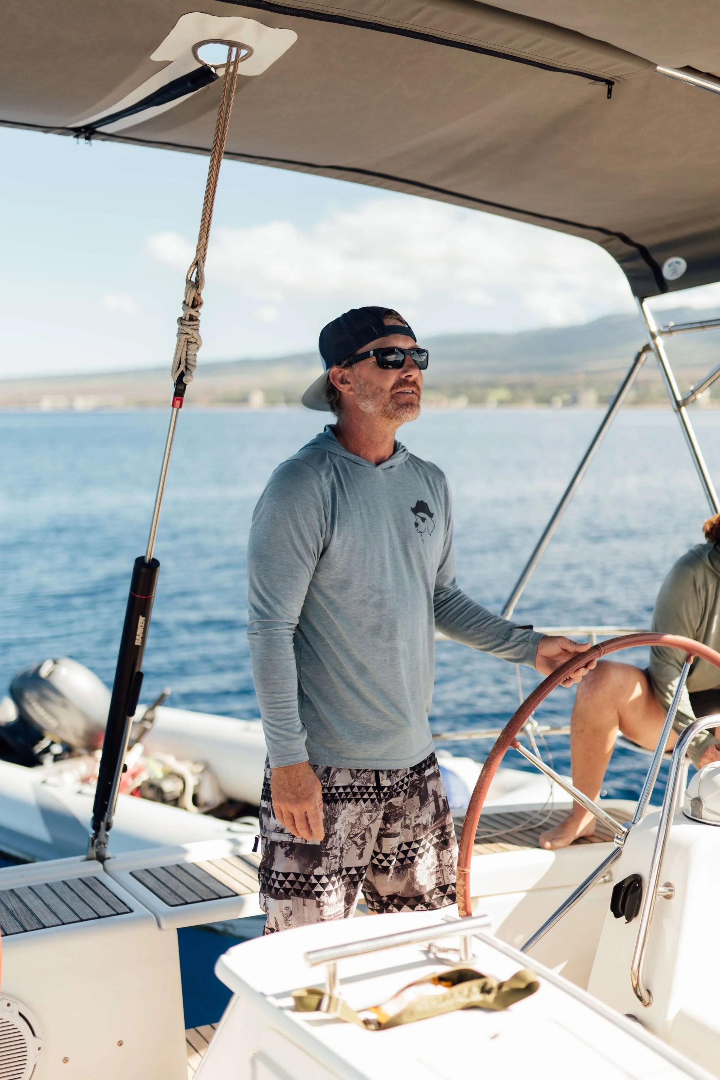 A man standing on a boat holding the steering wheel, wearing a gray long-sleeve shirt, patterned shorts, sunglasses, and a backwards baseball cap, with water and distant shoreline in the background.