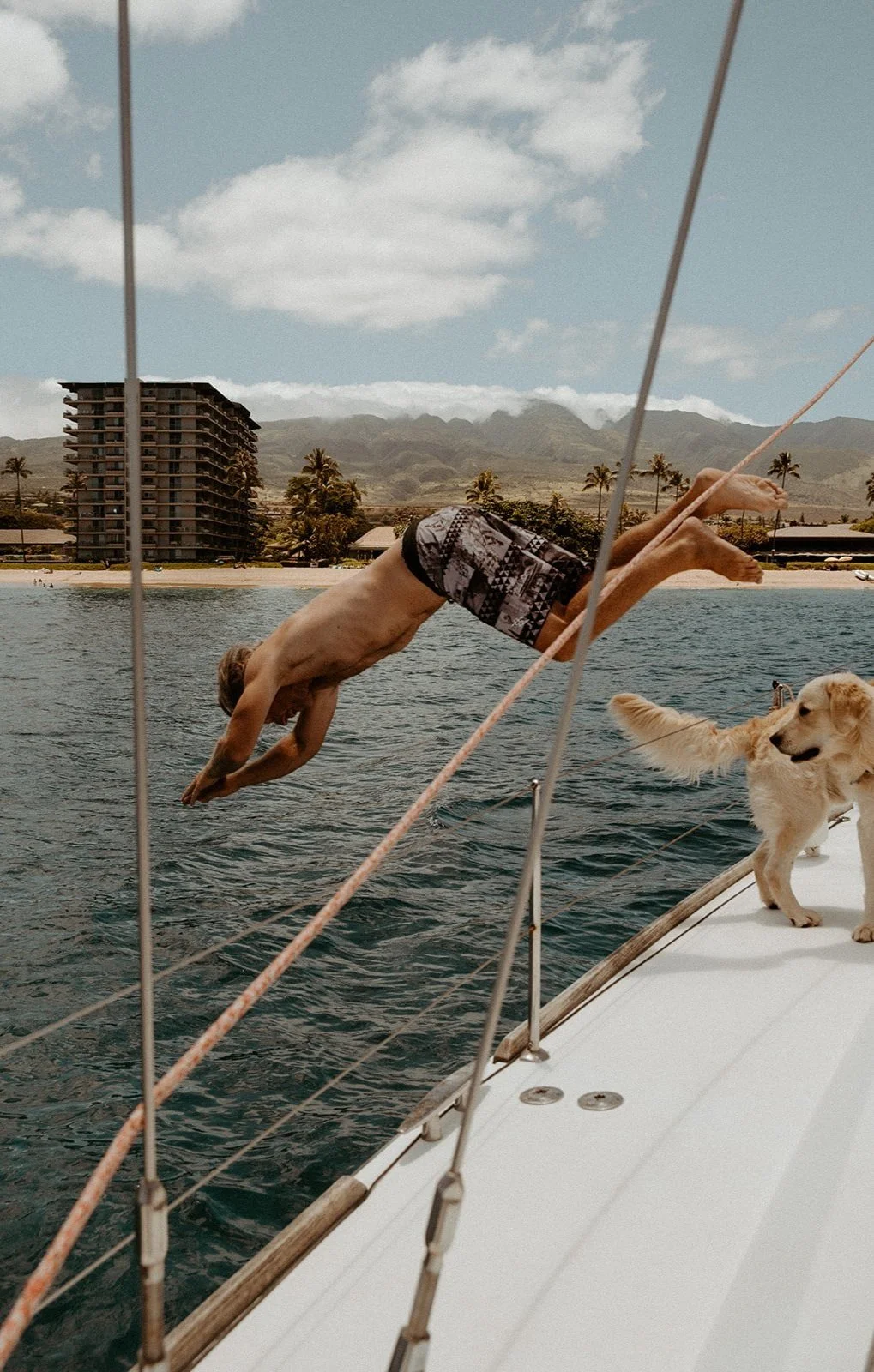 A man diving off a boat into the water with a dog on the deck, in a tropical landscape with mountains and a beach.