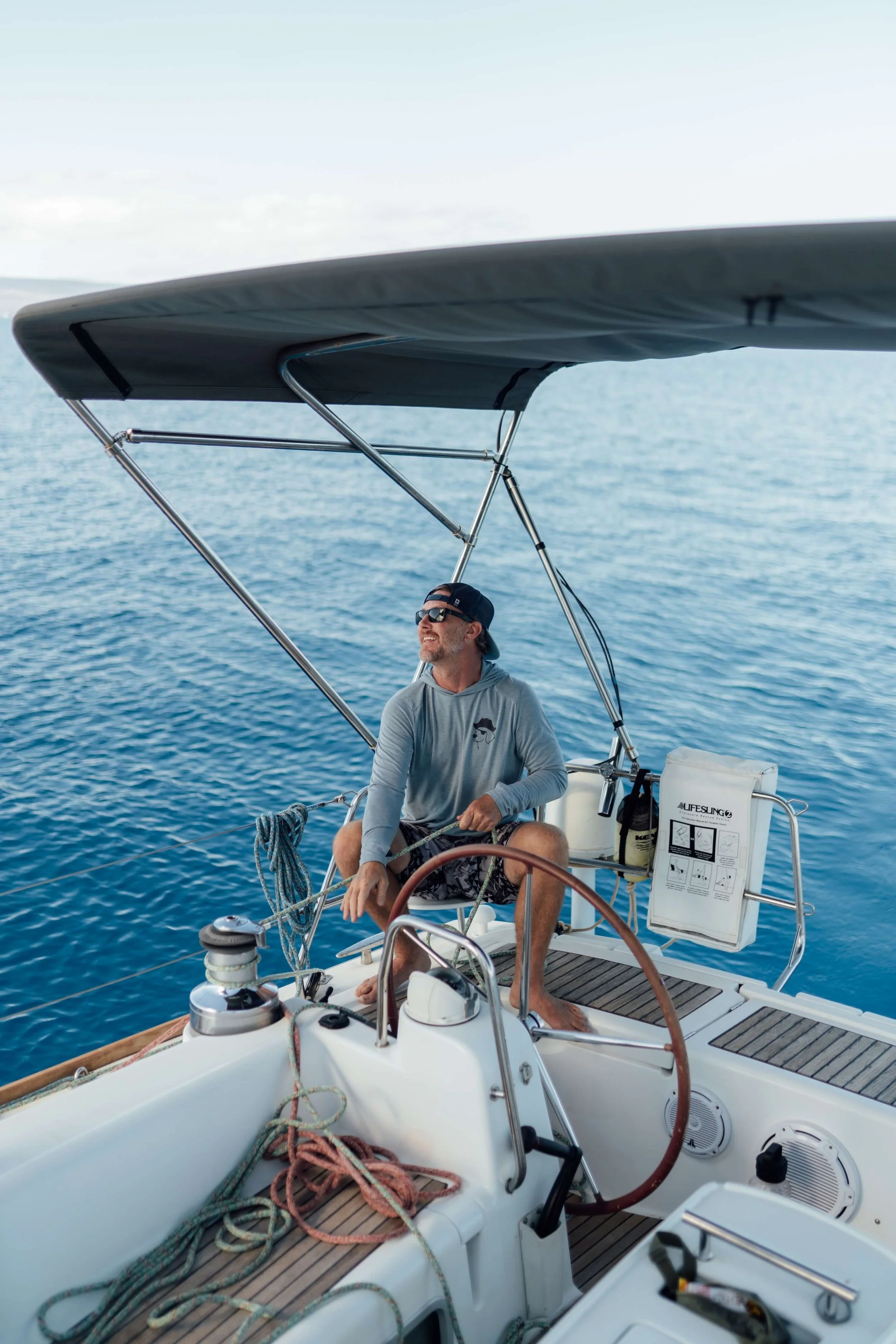 A man sitting on a sailboat in the ocean, wearing sunglasses and a hoodie, enjoying sunny weather under a shaded canopy.