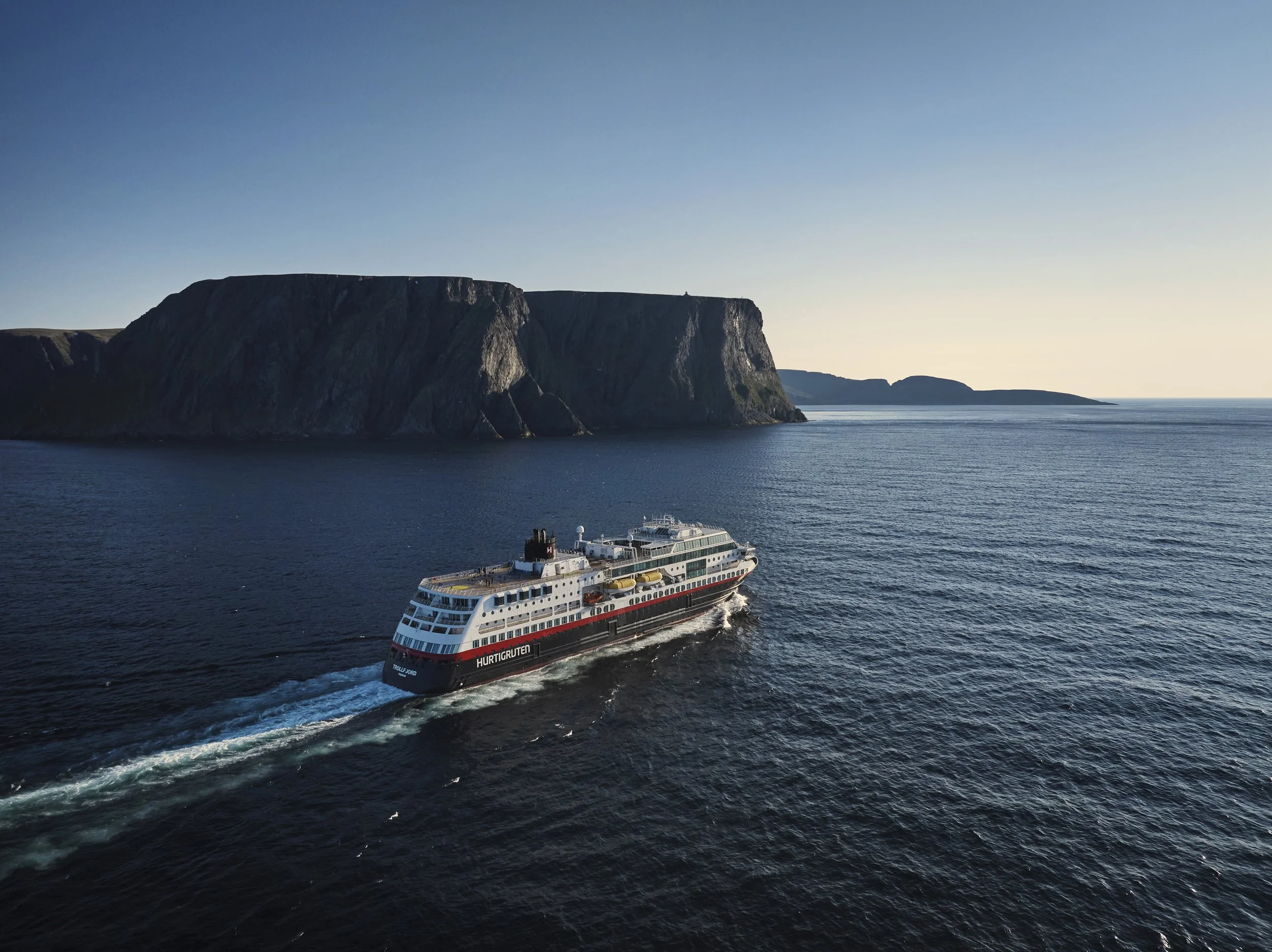 A cruise ship sailing near a large rocky island with cliffs, on calm ocean water during sunset or sunrise.