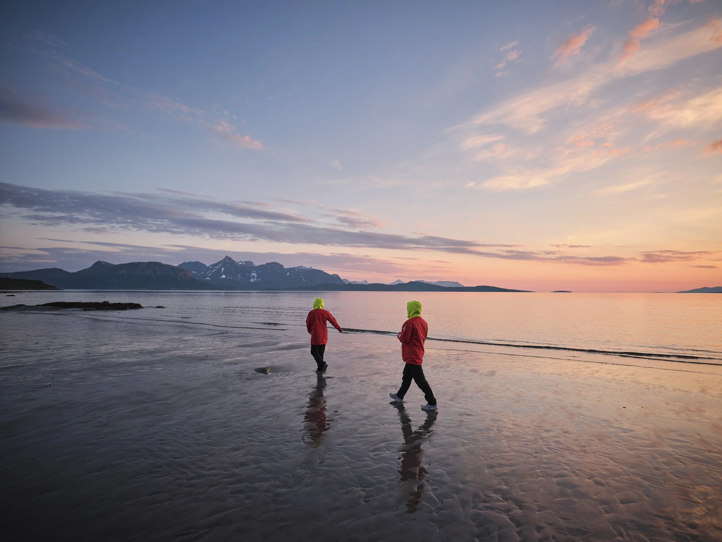 Two children wearing red jackets and green hats walking along a wet beach at sunset with a mountain range in the background.