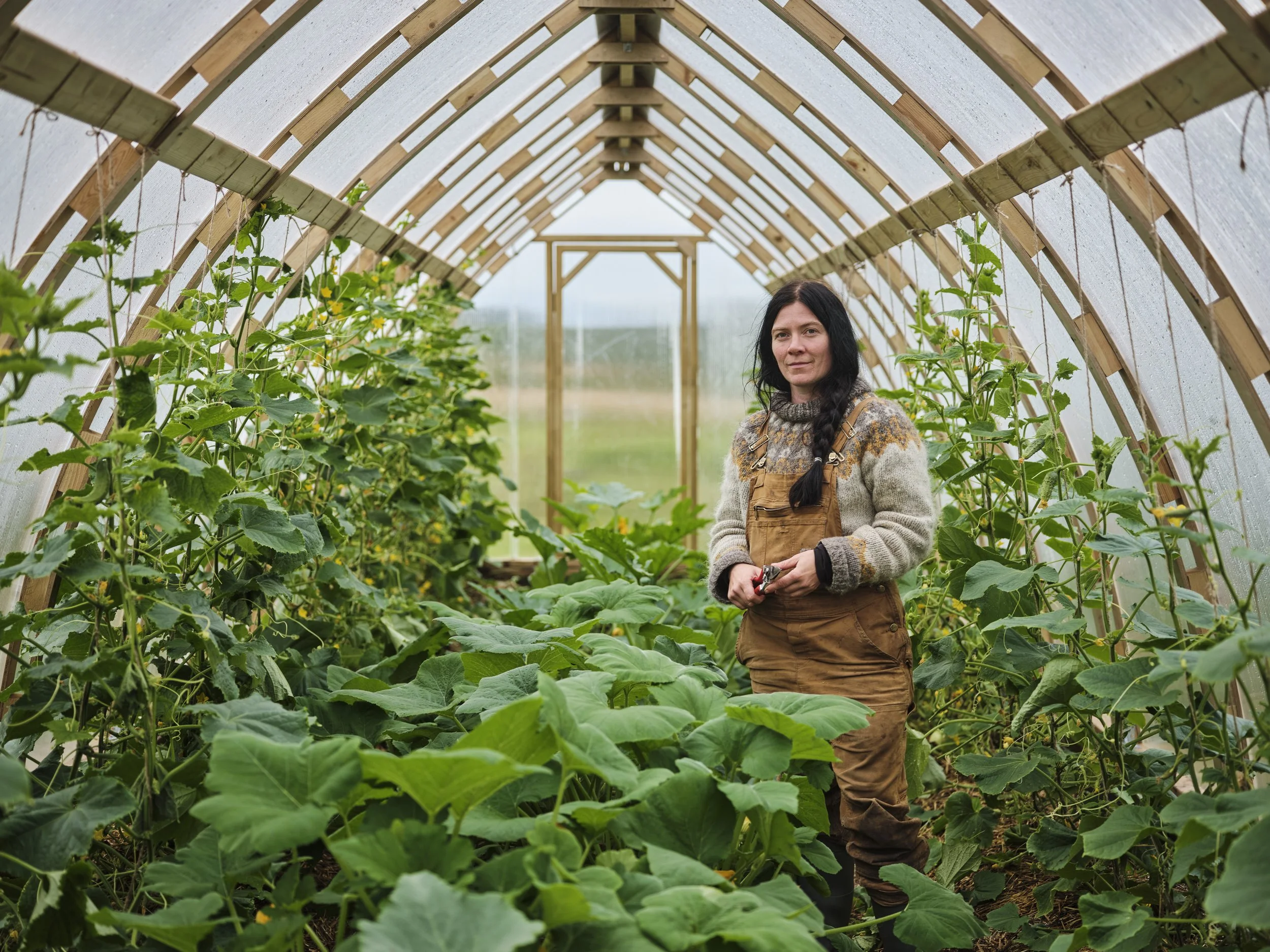 A woman with dark hair in a braid, wearing a sweater and brown overalls, standing inside a greenhouse with lush green plants.