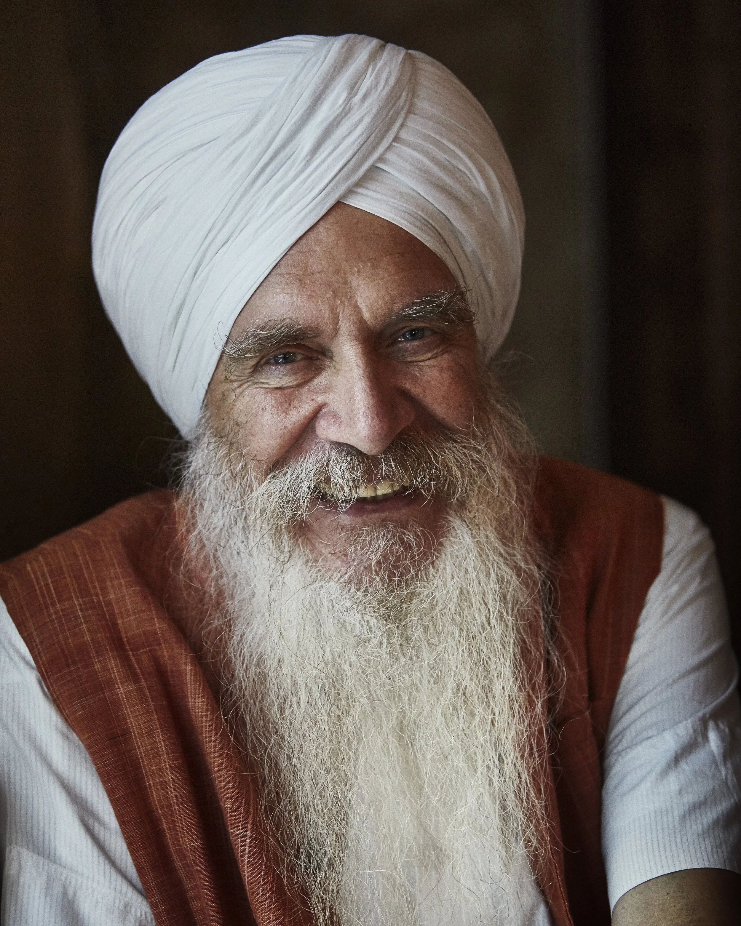 A smiling elderly man with a long white beard wearing a white turban and traditional attire.