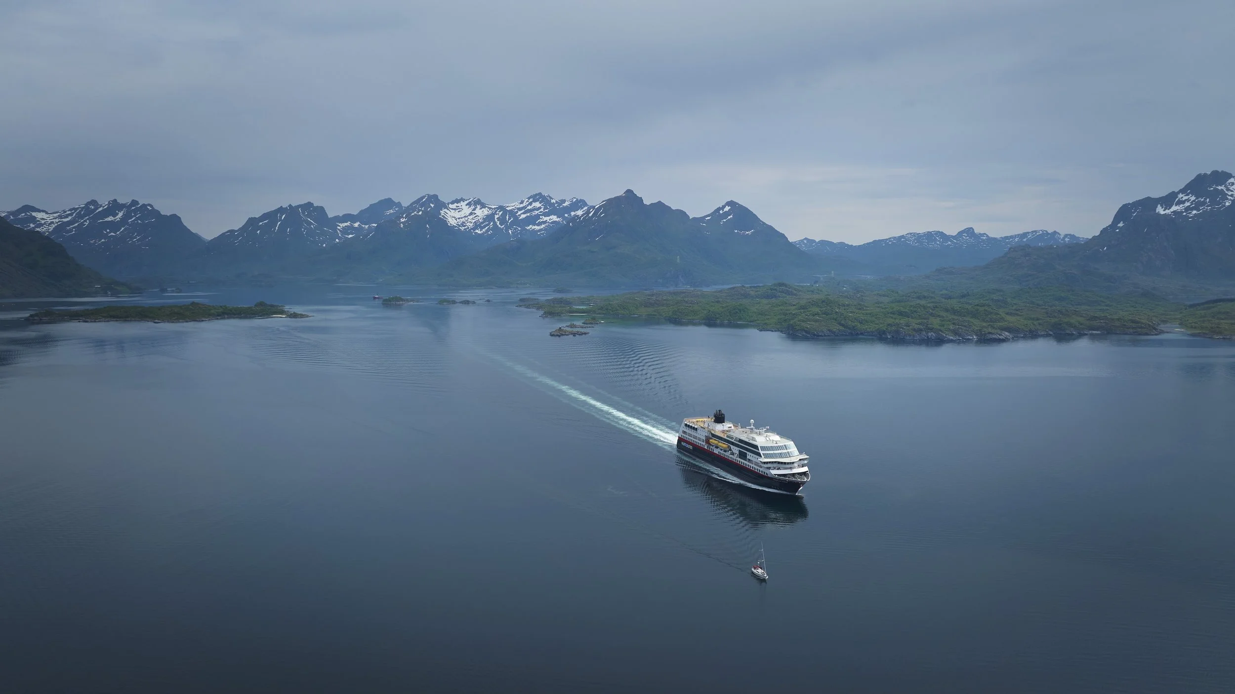 A large cruise ship sailing in a calm blue fjord with mountains, some snow-capped, in the background and smaller boats nearby.