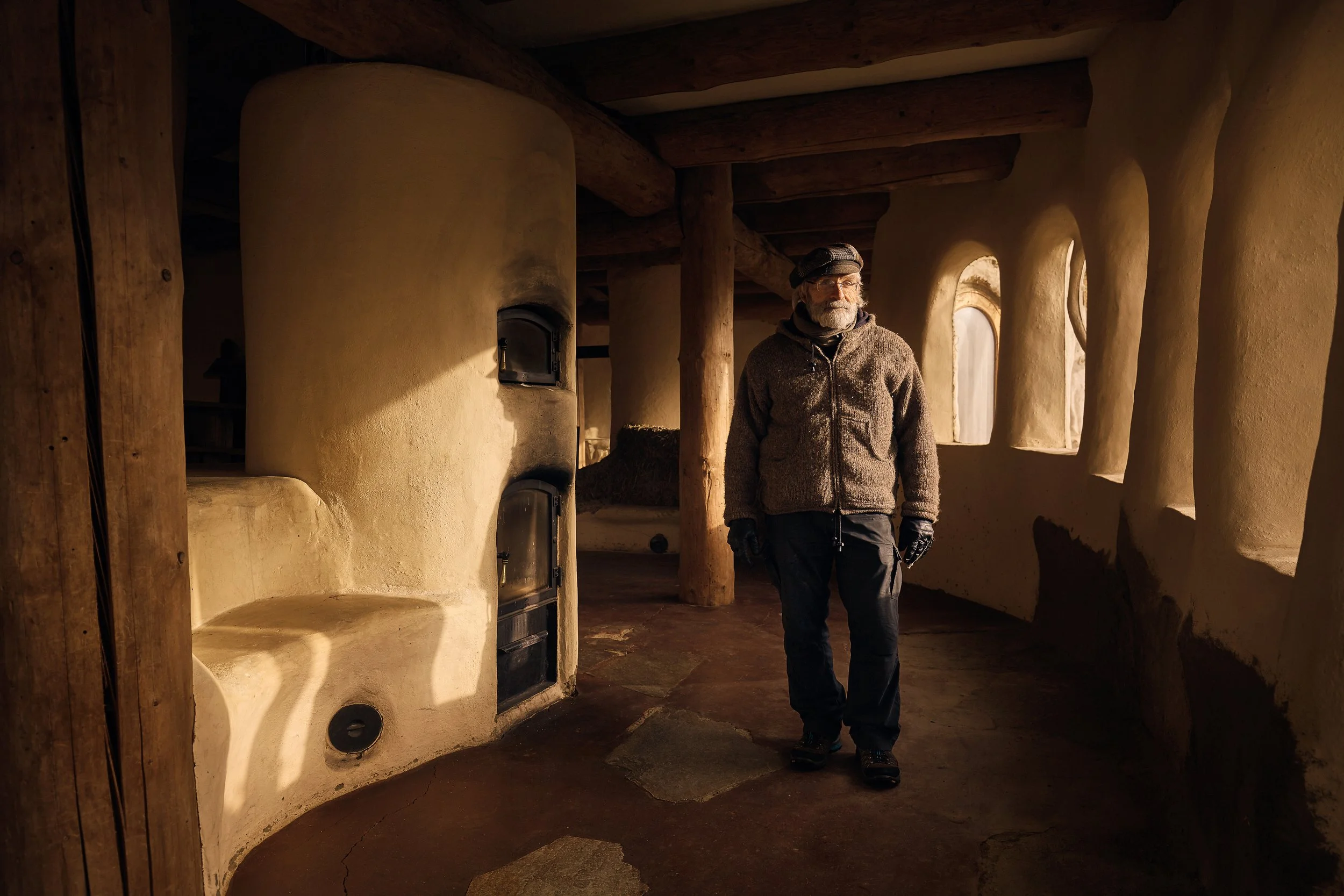 An elderly man wearing a brown fleece jacket, black gloves, and a beanie walking inside a house with earth-toned walls and wooden beams. Environmental portrait. Editorial.